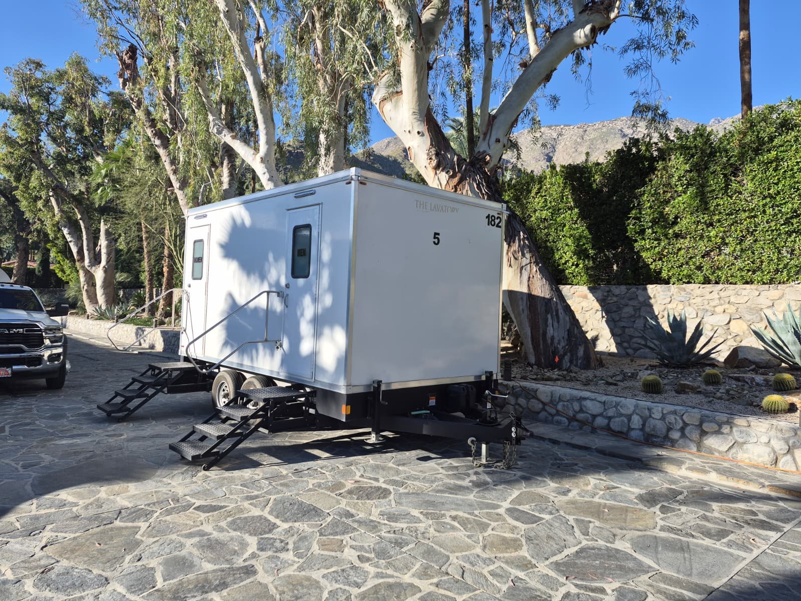 A Lavatory 5-Station luxury restroom trailer parked on a stone driveway under eucalyptus trees, with desert mountains behind it