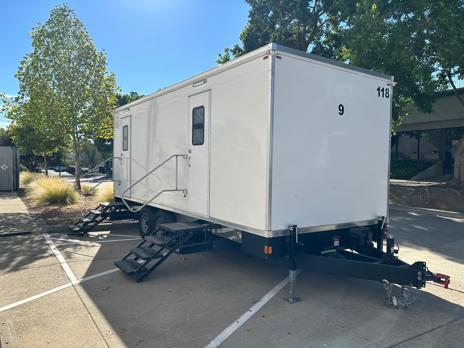 A Lavatory 9-Station restroom trailer parked across spaces in an office parking lot, with trees and landscaping behind it