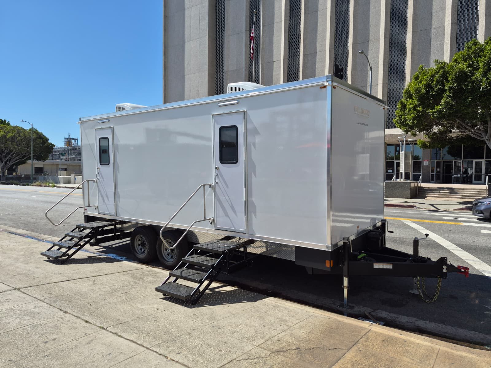 A long Lavatory restroom trailer parked along the curb in front of a tall office building on a sunny day