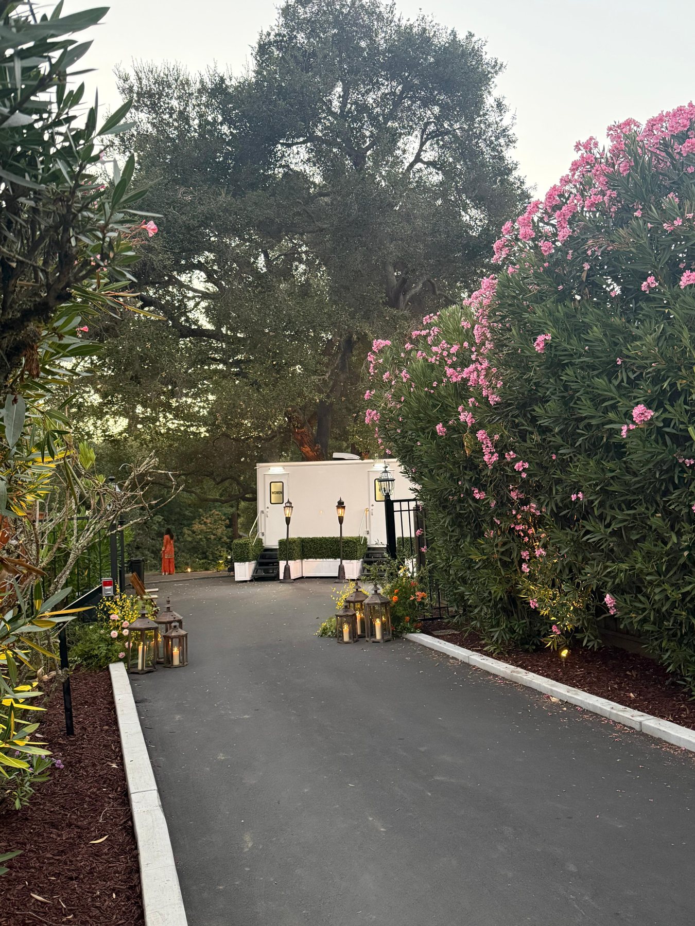 A Lavatory restroom trailer framed by hedge planters and lamp posts at the end of a lantern lined drive at a garden wedding venue