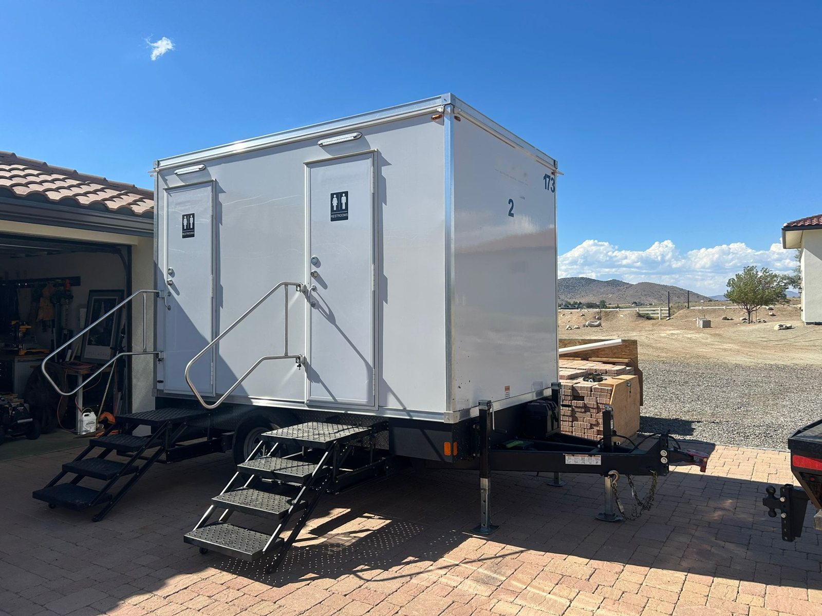 A two station Lavatory restroom trailer parked on a paver driveway beside an open garage, with stacks of pavers for a home project next to it