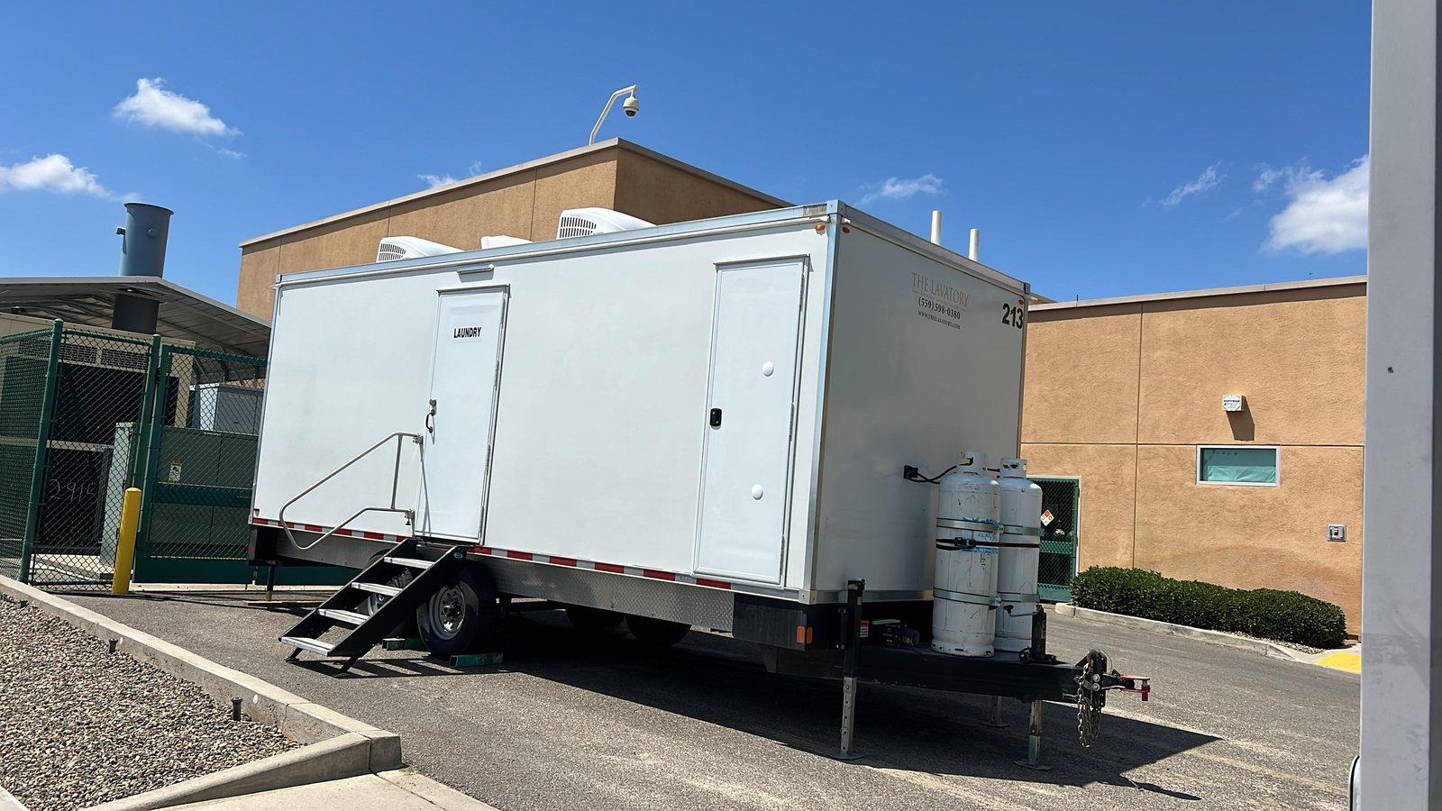 A Lavatory 8-Station laundry trailer with steel stairs and two upright propane tanks on the tongue, parked on a paved lot beside a stucco building