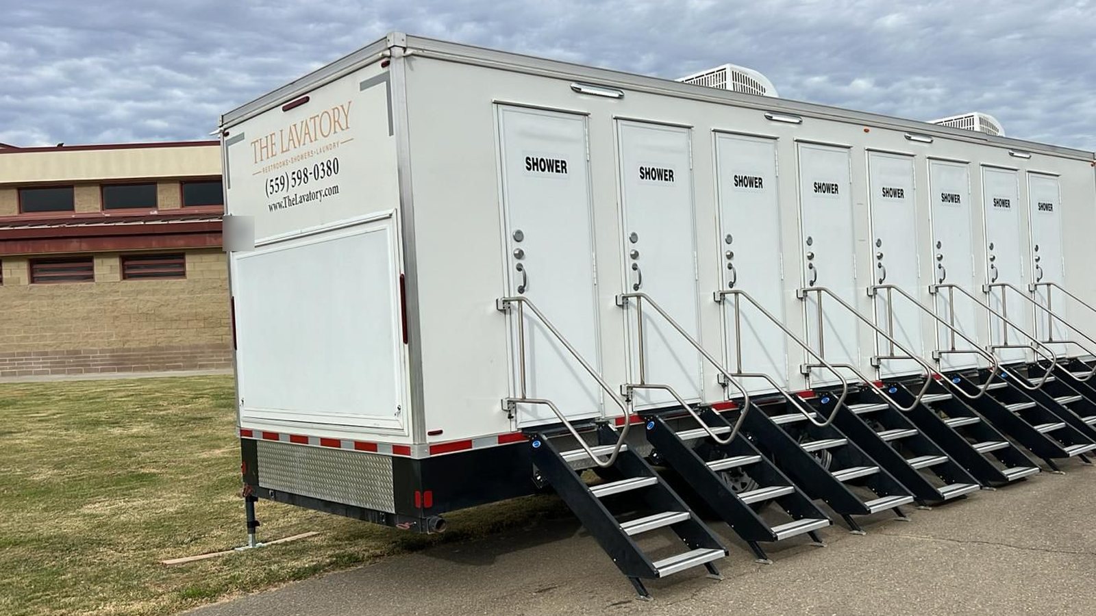 8-station shower trailer with a stair at each of its eight doors, parked on a paved drive beside a tan block building and a lawn