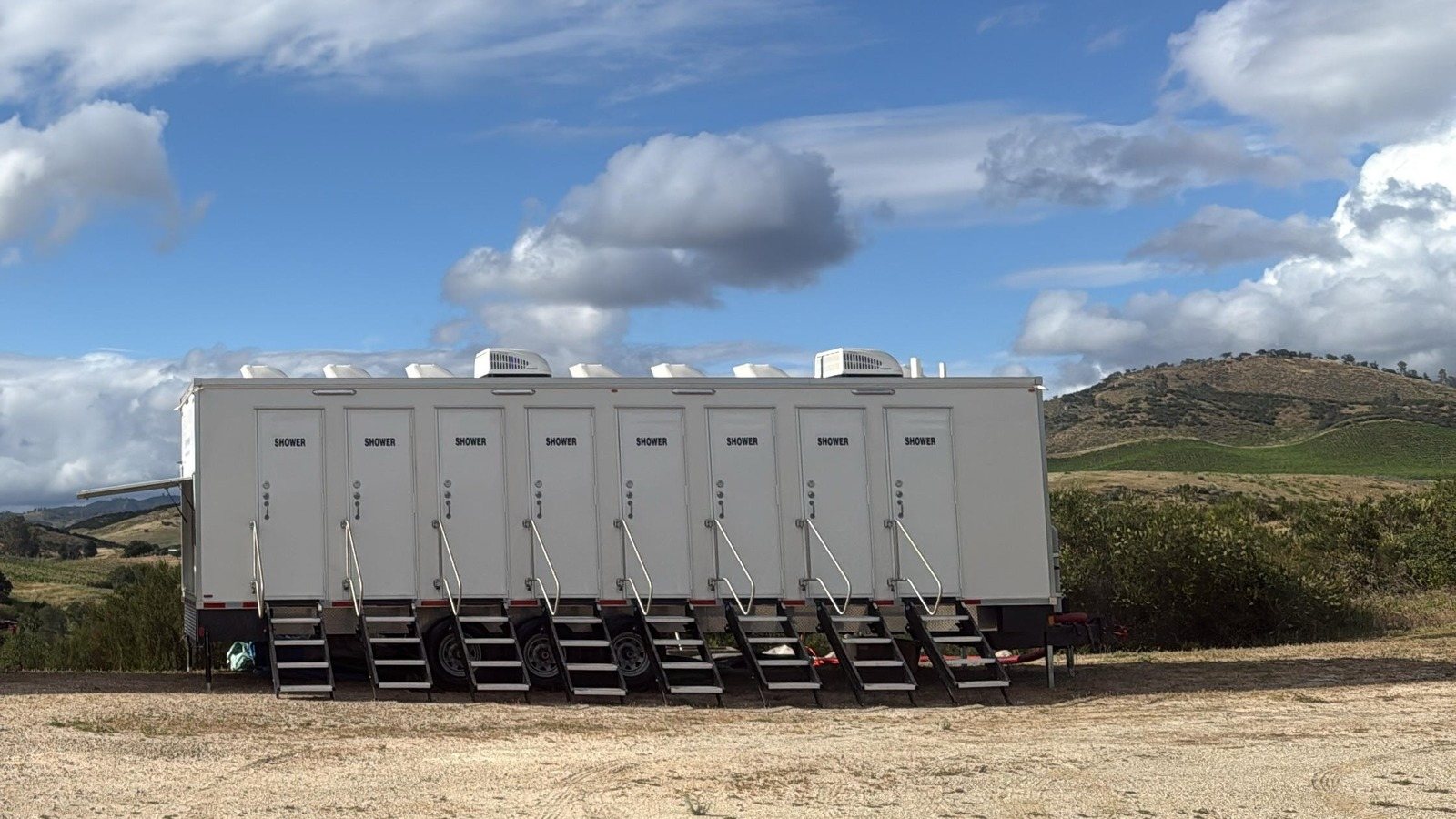8-station shower trailer with a stair at each of its eight doors, set on a dry dirt pad with vineyard rows and brown grassy hills behind