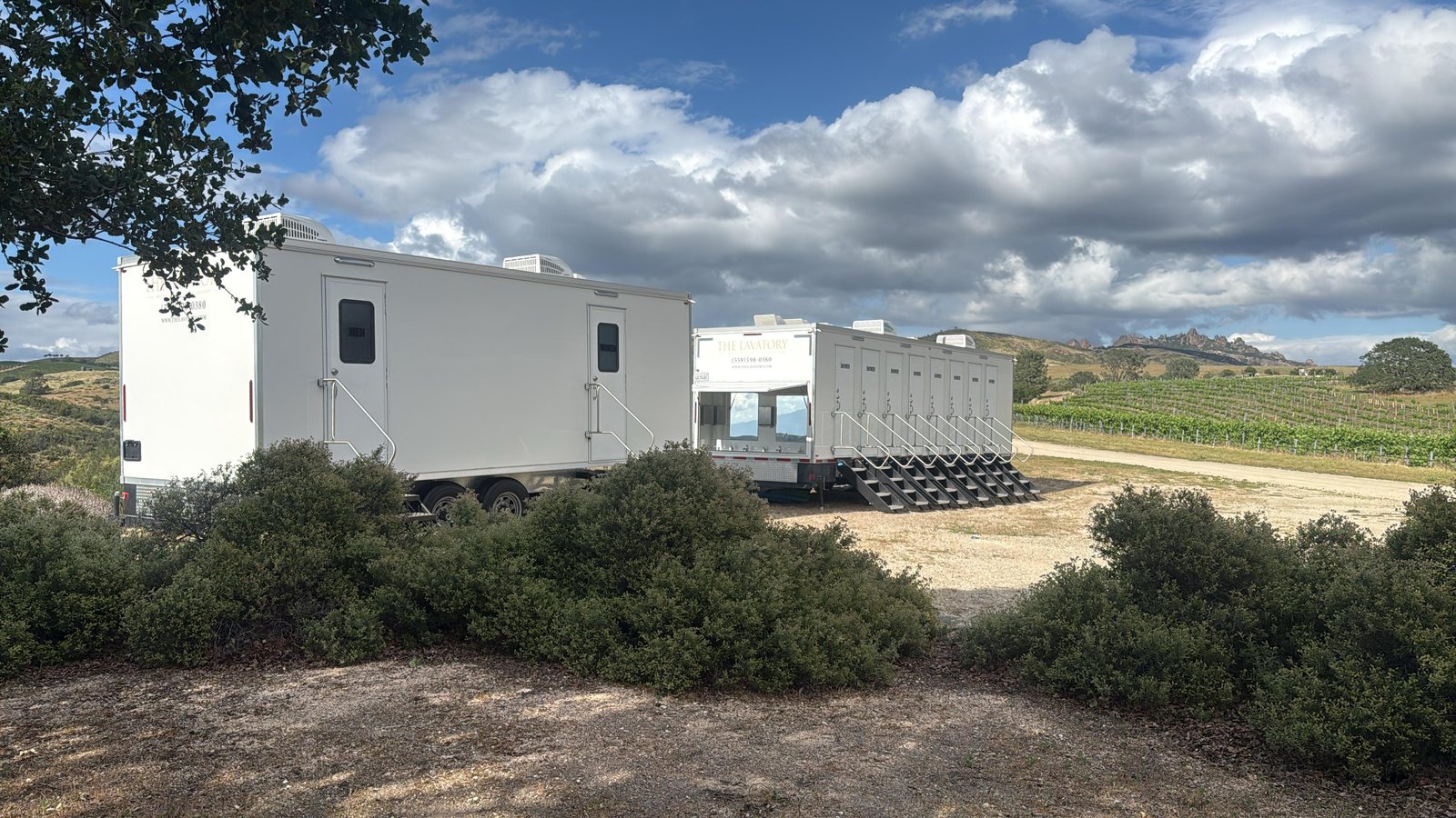 Restroom trailer and an 8-station shower trailer with its sink side open, parked on a dirt pad behind oak scrub with vineyard rows on the hills beyond
