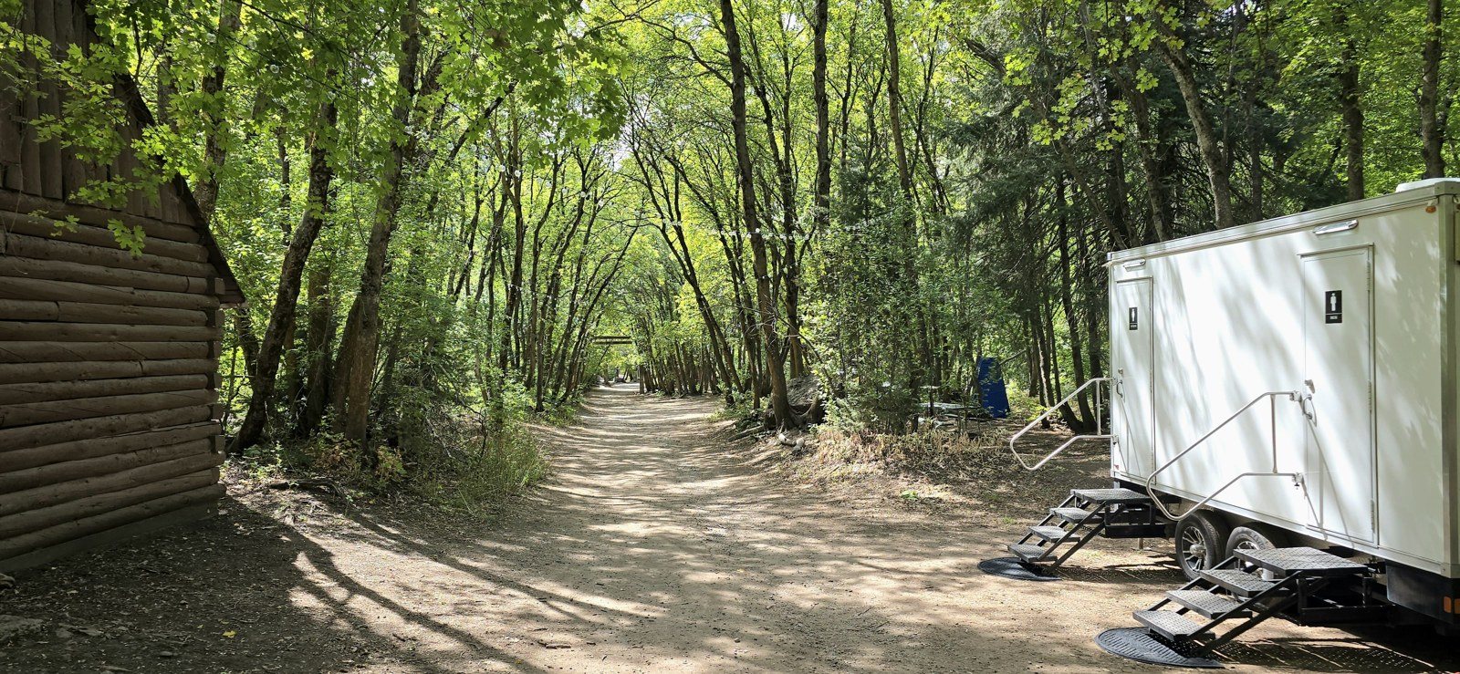 A Lavatory restroom trailer parked on a dirt lane beside a log cabin under a canopy of tall canyon trees