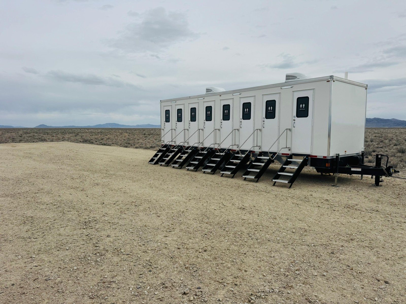 A long multi-station Lavatory restroom trailer set up for a work crew on a gravel pad at a remote high plains job site, with low mountains on the horizon