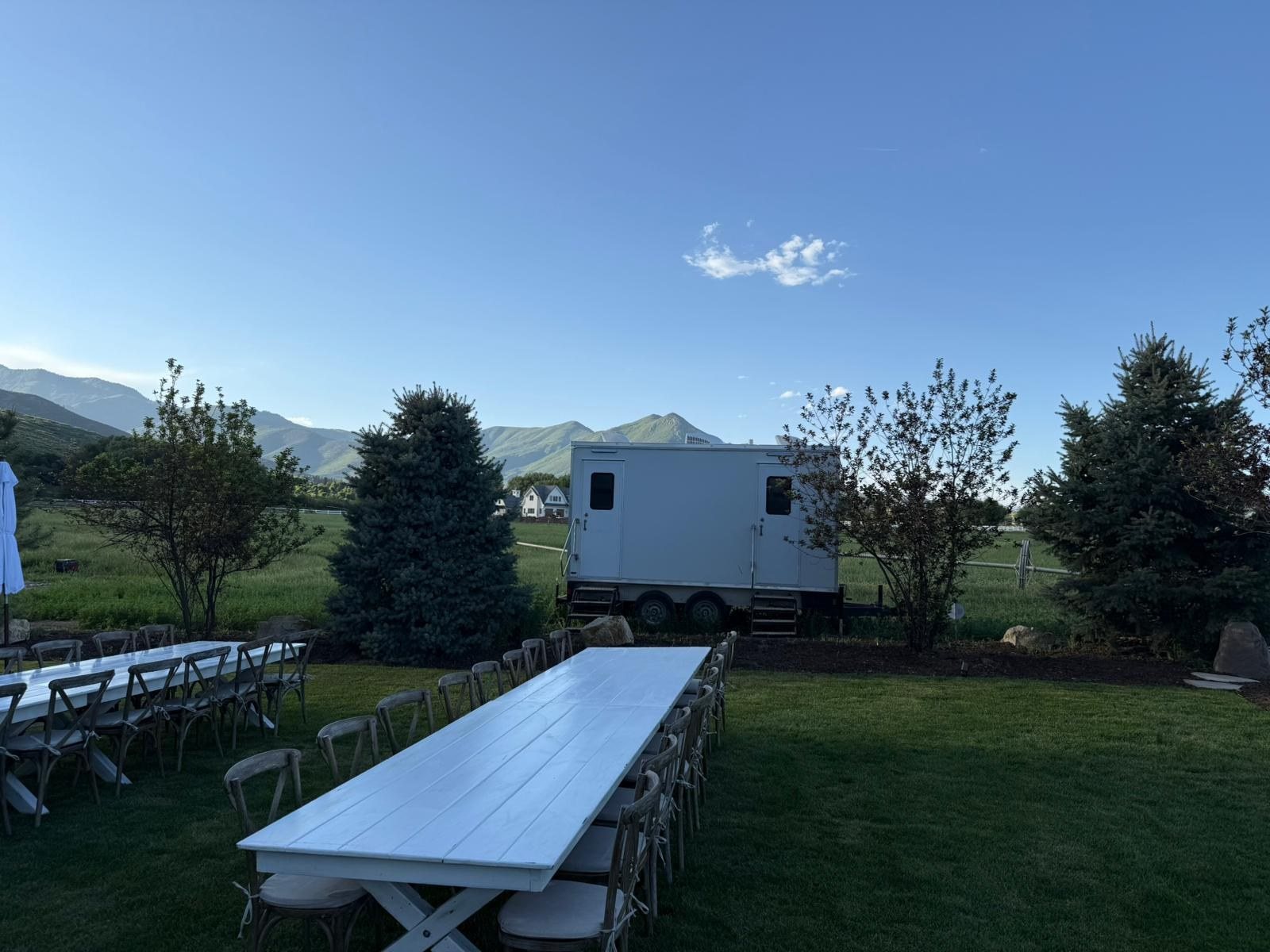 A Lavatory restroom trailer tucked behind blue spruce trees at a ranch wedding, with long white farm tables set on the lawn and mountains behind