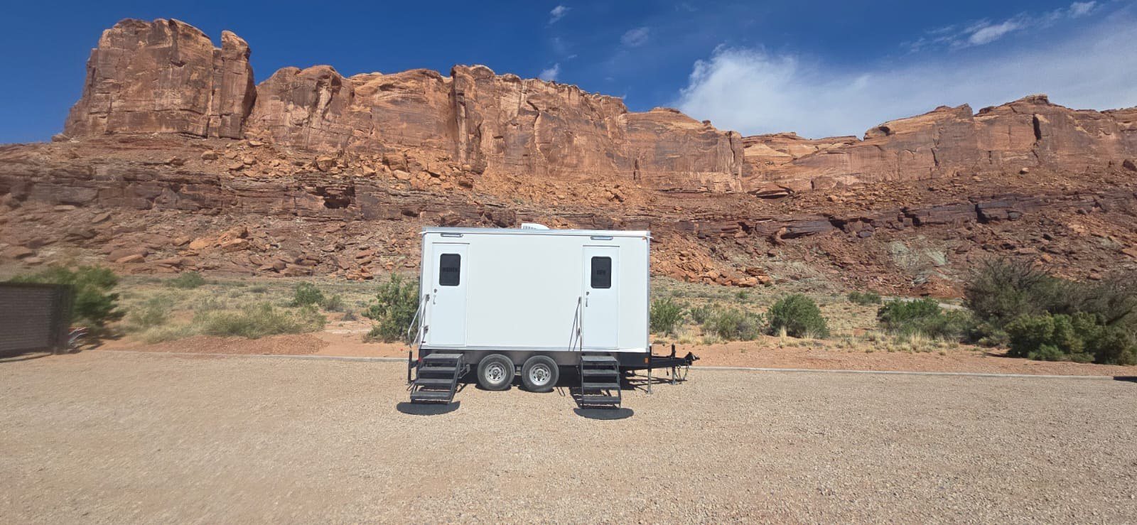 A Lavatory two-room restroom trailer leveled on a gravel pad below red sandstone cliffs
