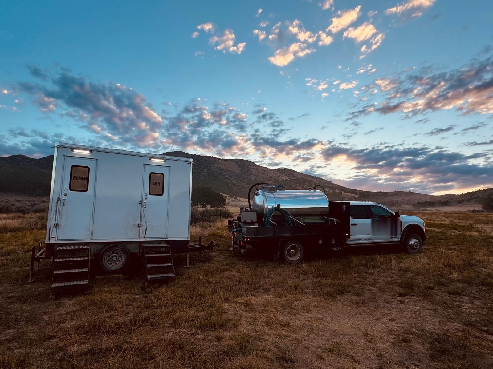 A Lavatory service crew truck with a fresh water and waste tank parked next to a two-room restroom trailer in a mountain meadow at dusk