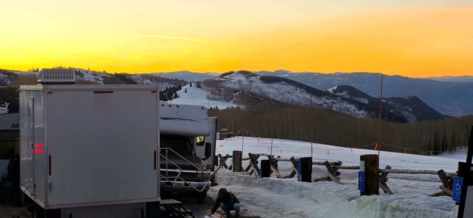 A Lavatory restroom trailer at a snowy ski area at sunset while a crew member works beside it, with snow-covered ridges behind