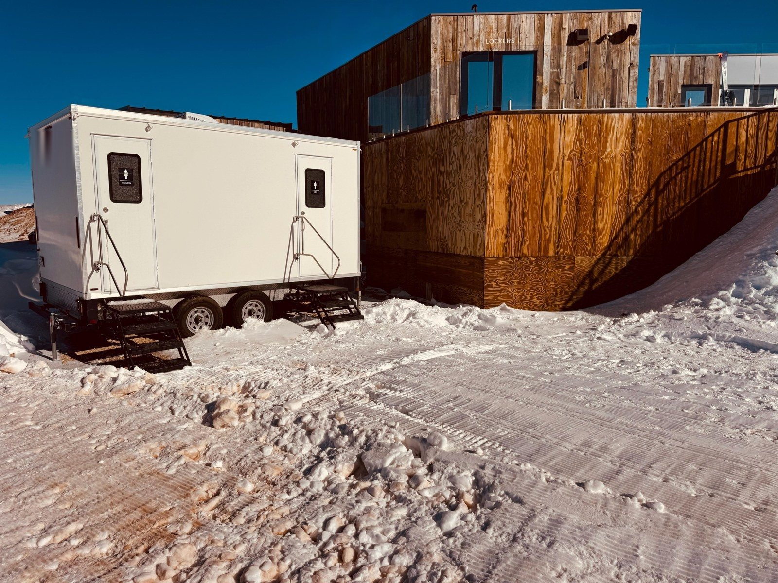 A Lavatory restroom trailer set on packed snow beside a timber-clad ski base building under a deep blue sky