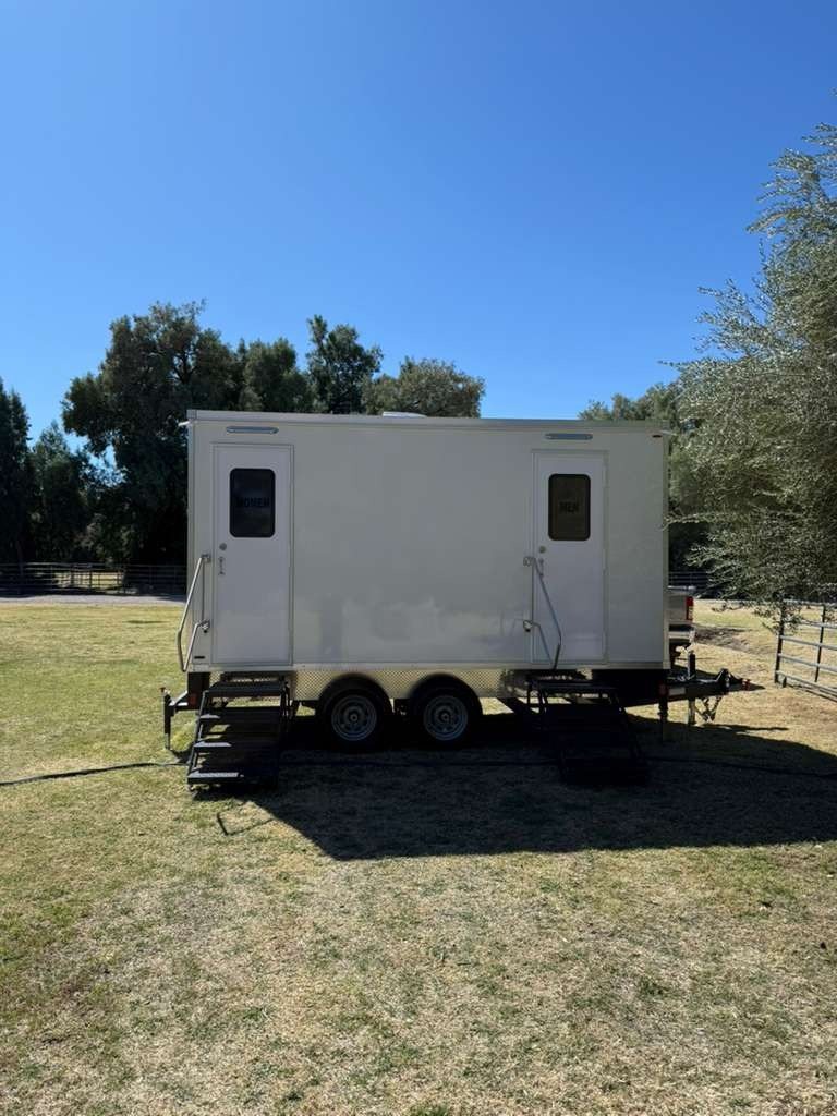 A Lavatory 2-Station restroom trailer with women's and men's doors parked on a dry grass lawn at a ranch, with tall trees and a pipe fence behind it