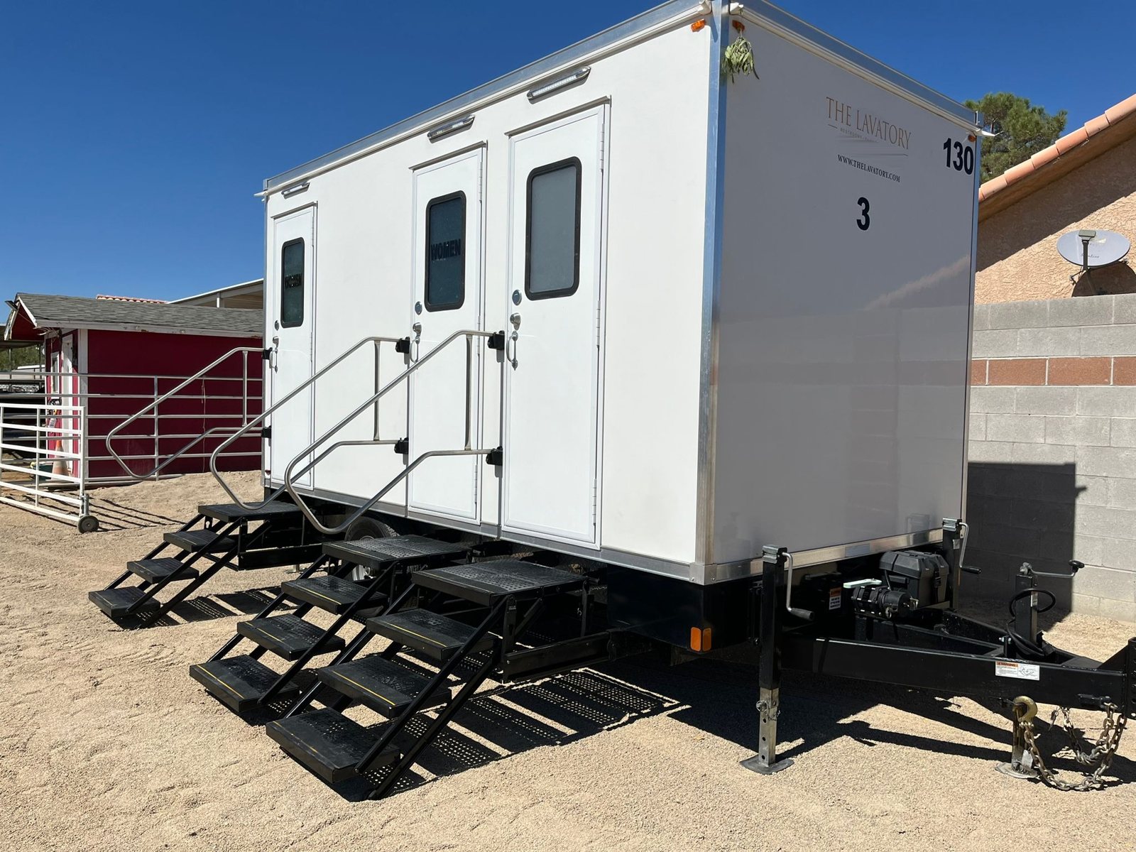 A Lavatory 3-Station restroom trailer with steel stairs at each door, set on packed desert dirt beside a block wall and a red barn