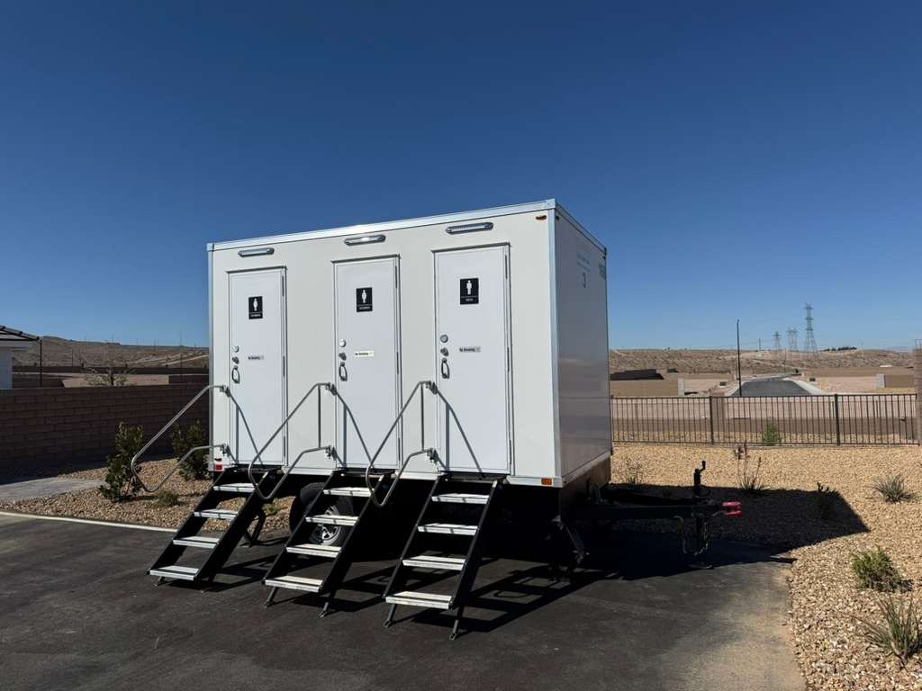 A Lavatory 3-Station restroom trailer with three steel stair sets on a paved lot beside desert landscaping, with power lines and bare hills behind a new subdivision
