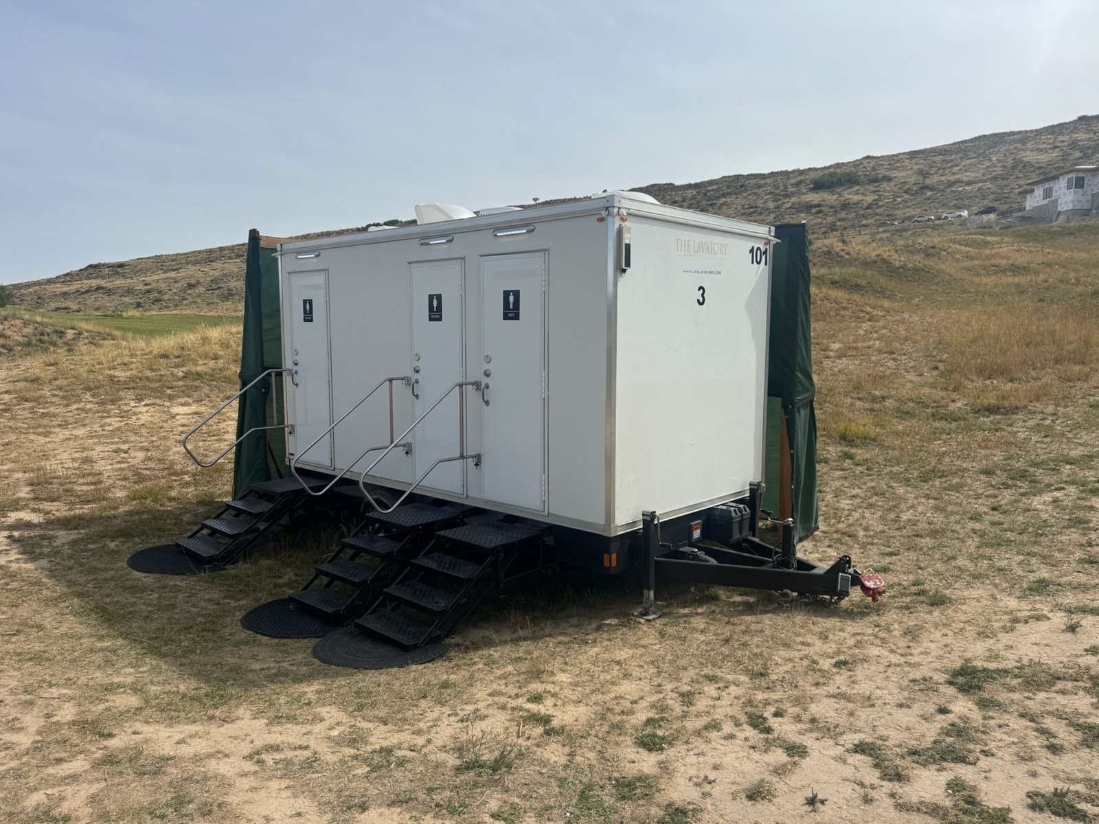A Lavatory 3-Station restroom trailer with three sets of steel stairs and green privacy screens on a dry grassy hillside event site