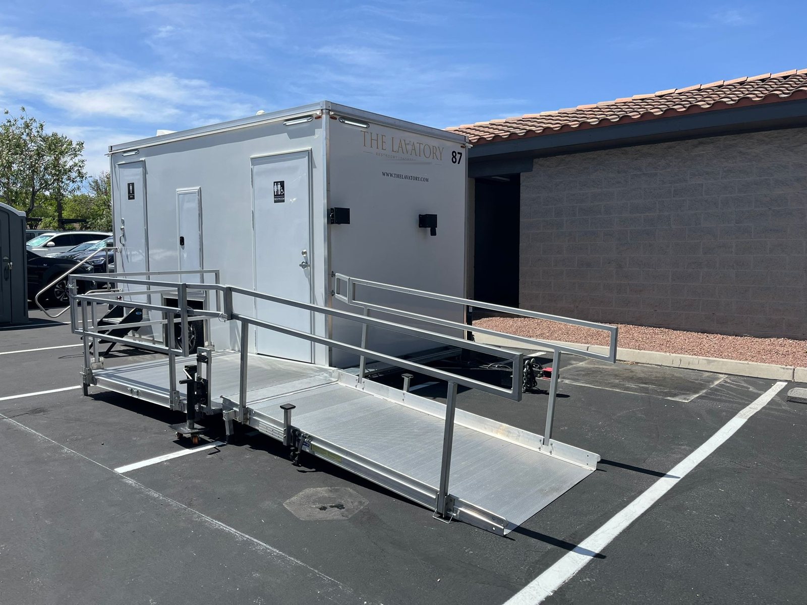 A Lavatory ADA + 2 restroom trailer with a long aluminum ramp and railings set in a parking lot beside a tile-roofed building and red gravel landscaping