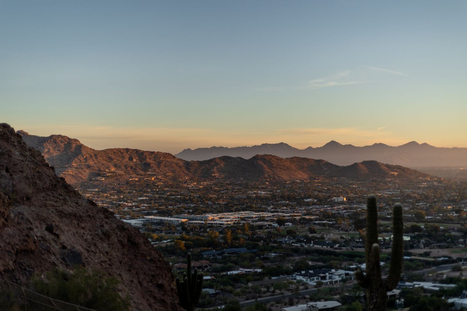 The Phoenix area at sunset seen from a desert peak, with saguaro cactus in the foreground and mountain ridges across the valley