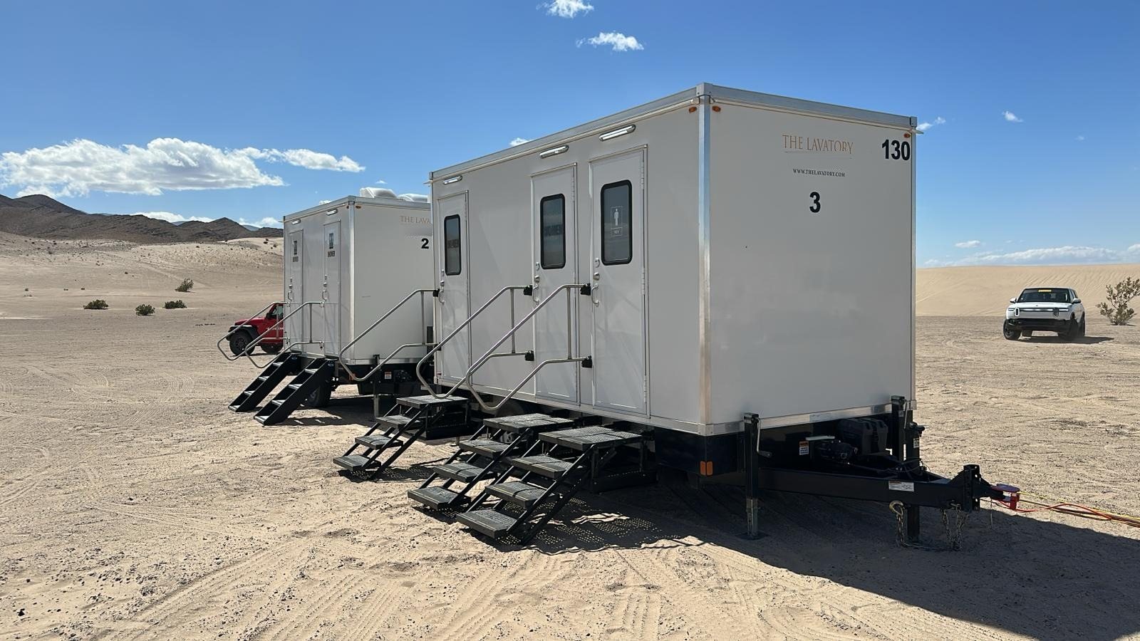 Two white Lavatory restroom trailers with steel stairs at every door set on open desert sand below bare mountains