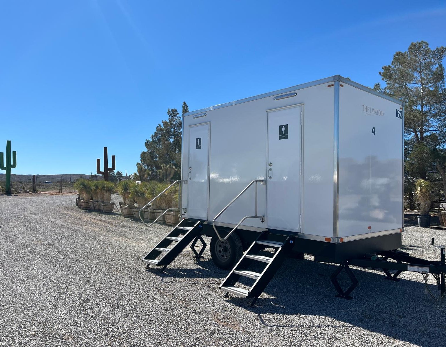 A Lavatory 2-Station restroom trailer with women's and men's doors and two sets of steel stairs, parked on a gravel lot beside desert landscaping with metal saguaro cactus sculptures and potted yuccas under a clear blue sky