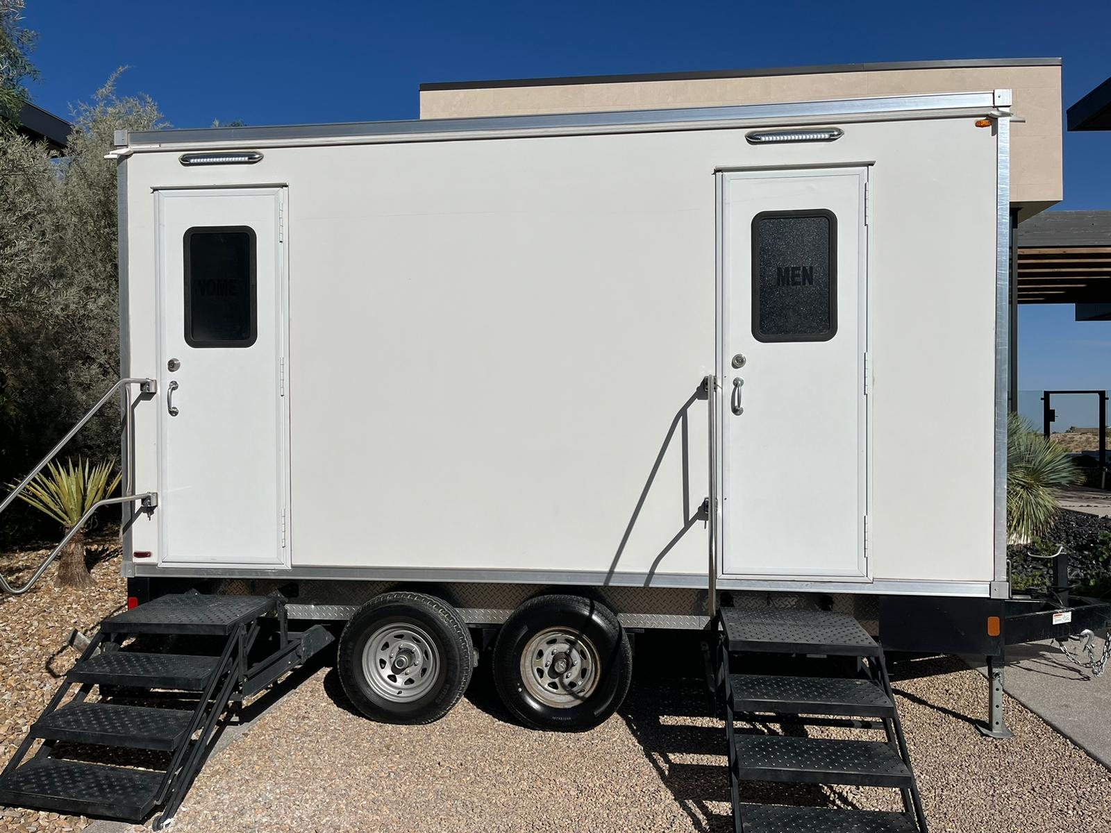 A clean white Lavatory 2-Station restroom trailer with women's and men's doors and black steel stairs, set on decomposed granite beside desert plants and a modern stucco home