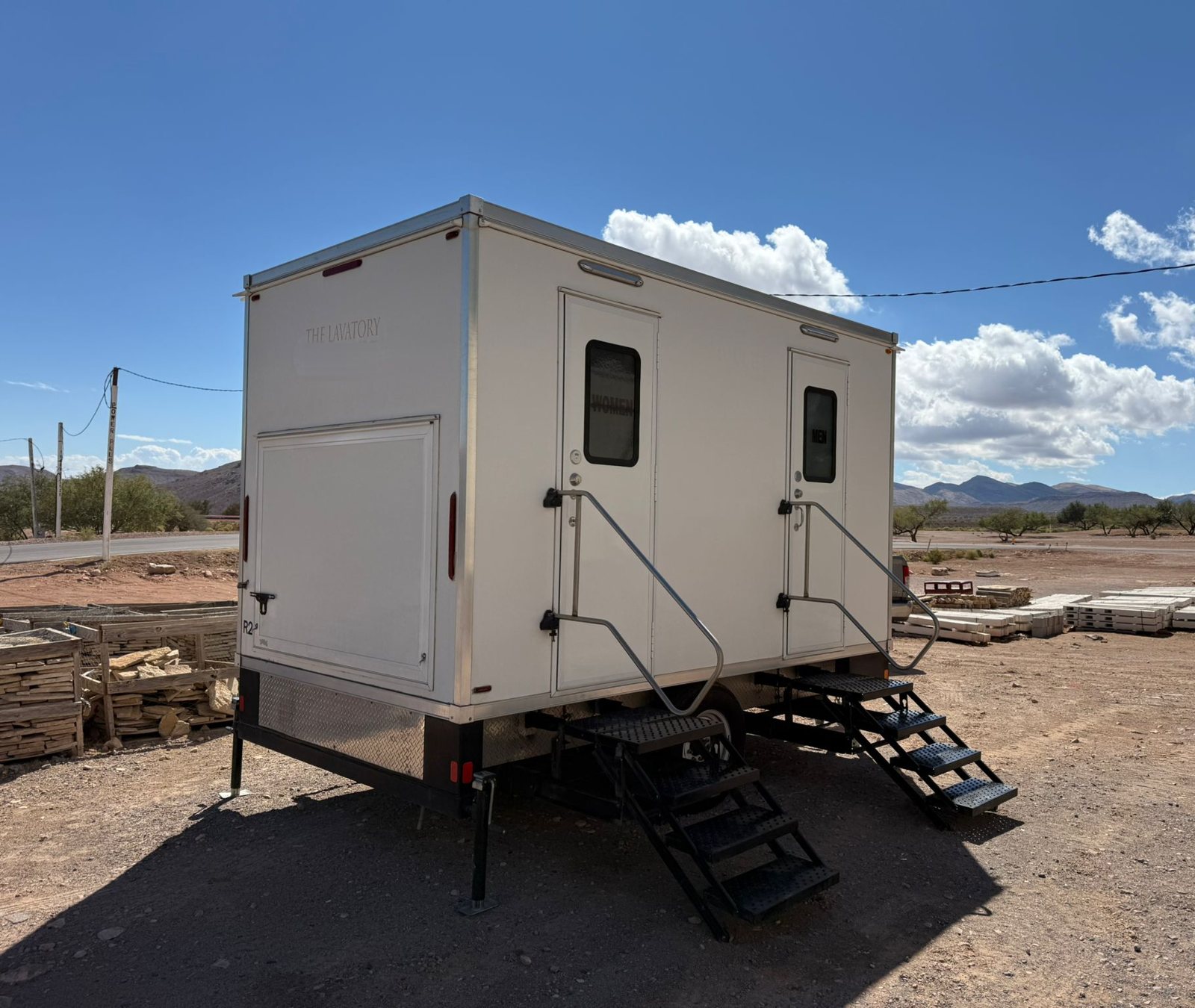 A Lavatory 2-Station restroom trailer with stairs at both doors, parked on a dirt lot with stacked stone pallets, mesquite trees and desert mountains behind it