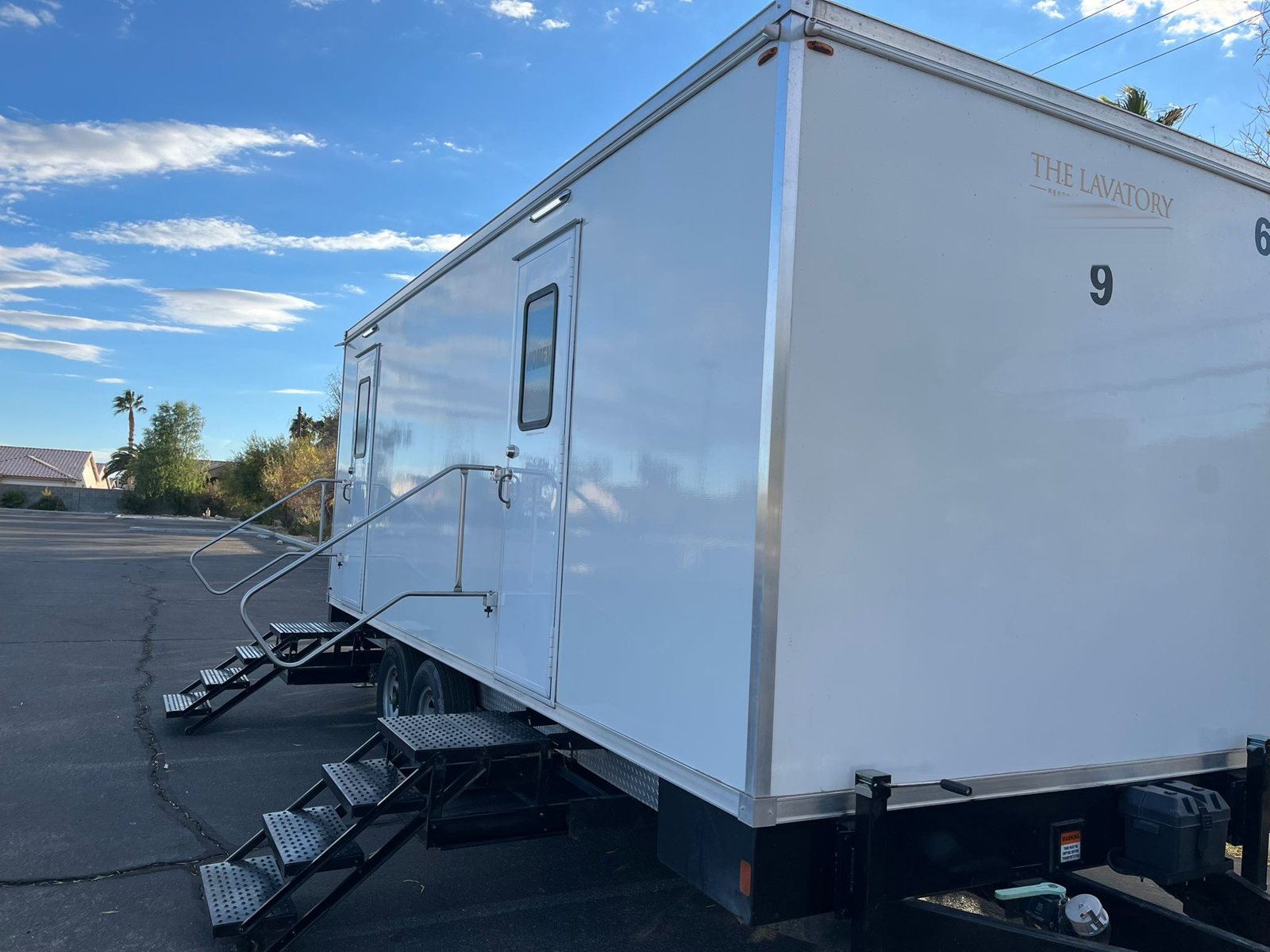A long Lavatory 9-Station restroom trailer with railed stairs, parked in a paved lot near tile-roof homes and palm trees on a sunny afternoon