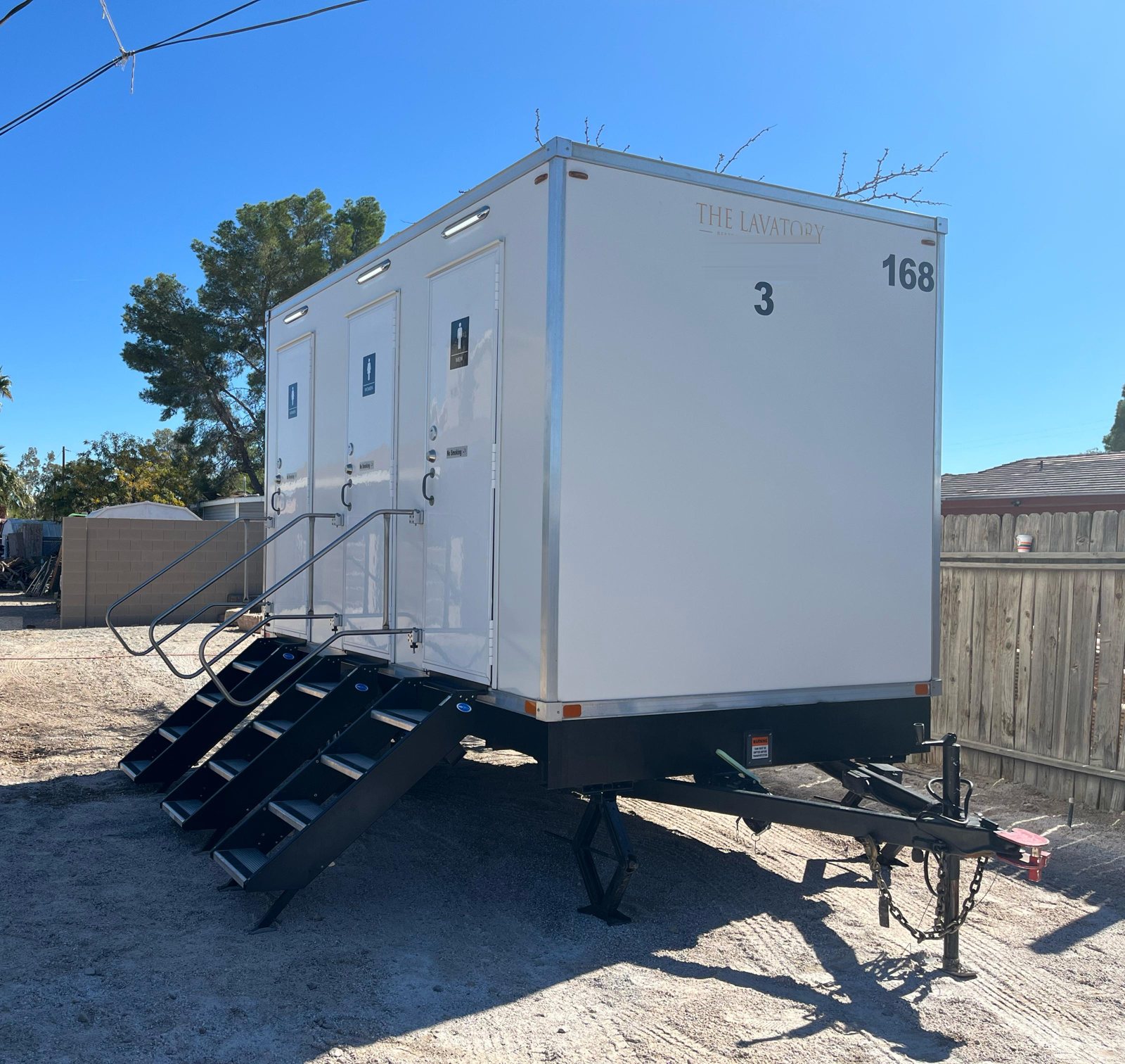 A Lavatory 3-Station restroom trailer with three doors and three sets of stairs, set on a gravel yard beside a block wall, a wood fence and tall palm trees
