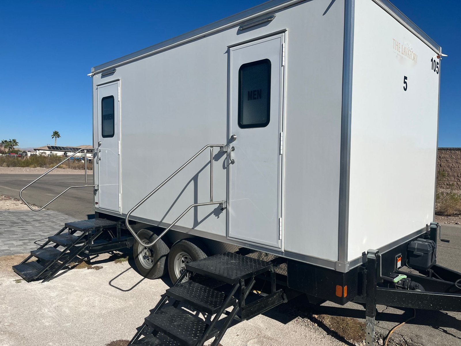 A Lavatory 5-Station restroom trailer with separate women's and men's doors and steel stairs, parked on a desert lot under a clear blue sky with palm trees behind it