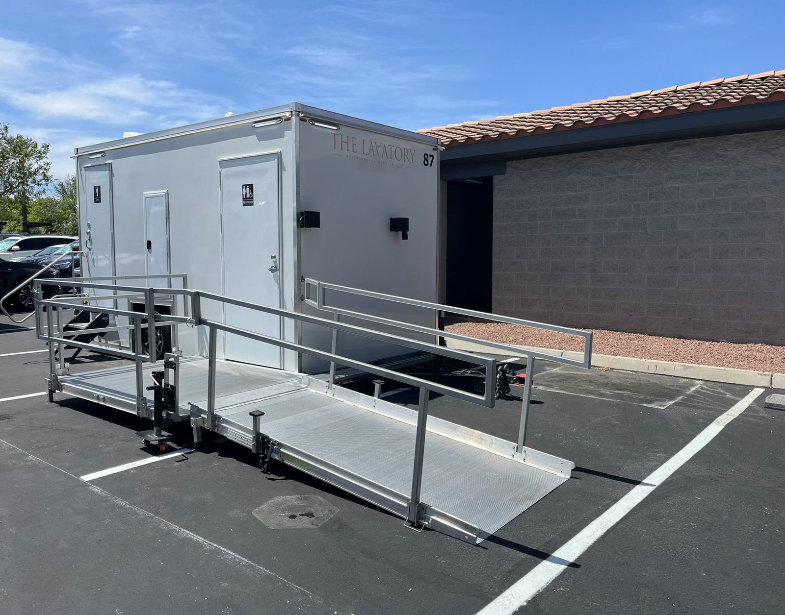 A Lavatory ADA restroom trailer with a long aluminum ramp and handrails set in a business parking lot next to a block building with a tile roof