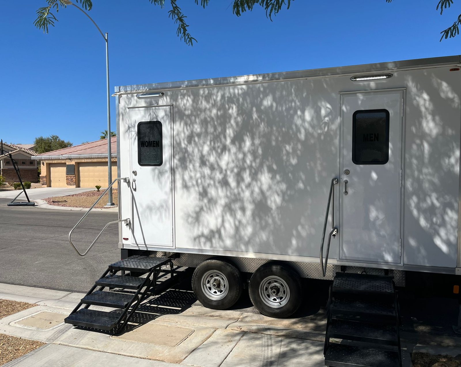A Lavatory 2-Station restroom trailer parked at the curb of a desert neighborhood street with stucco homes and tile roofs