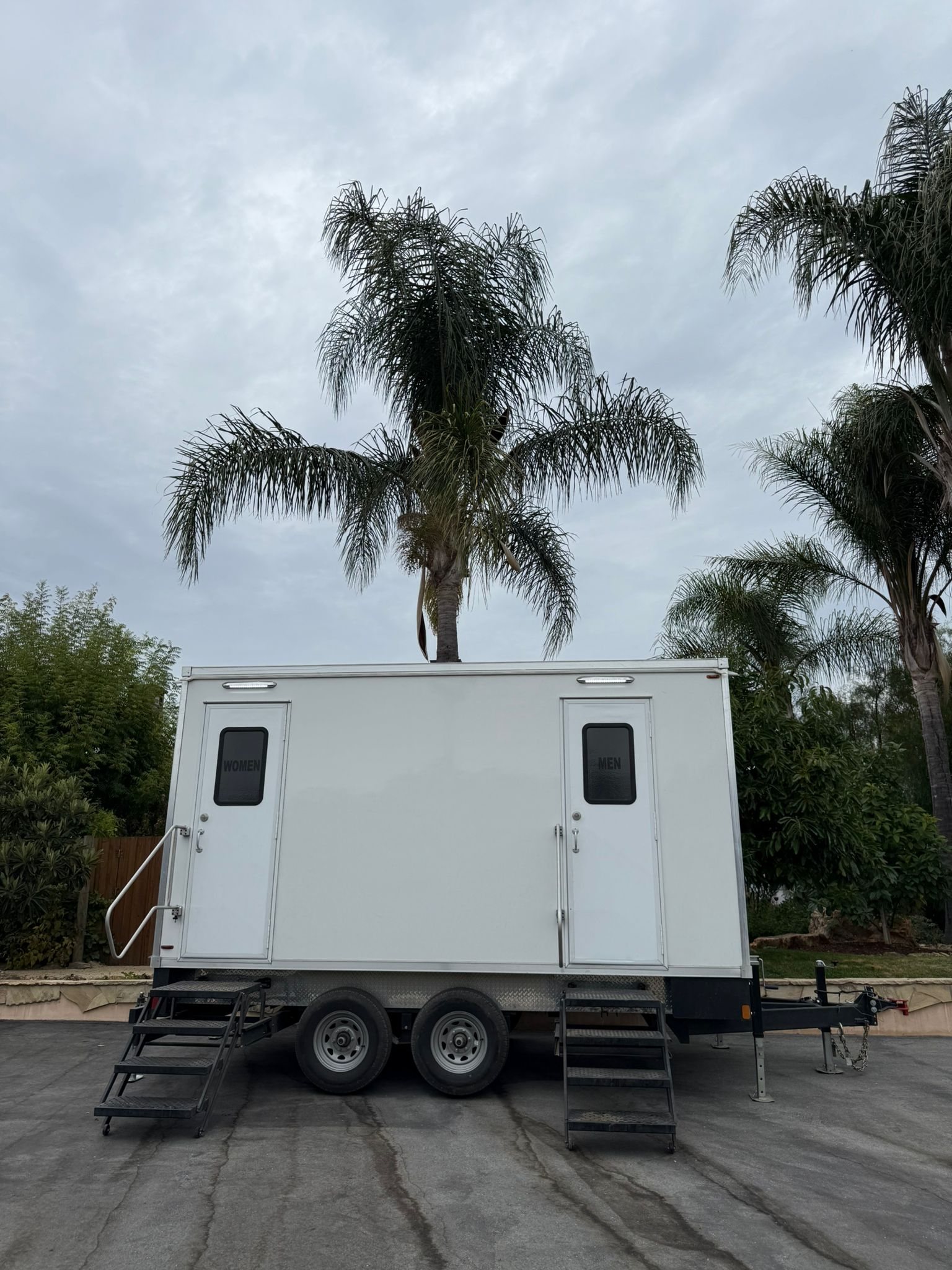 A white Lavatory 2-Station restroom trailer with women's and men's doors parked on a paved driveway under tall palm trees