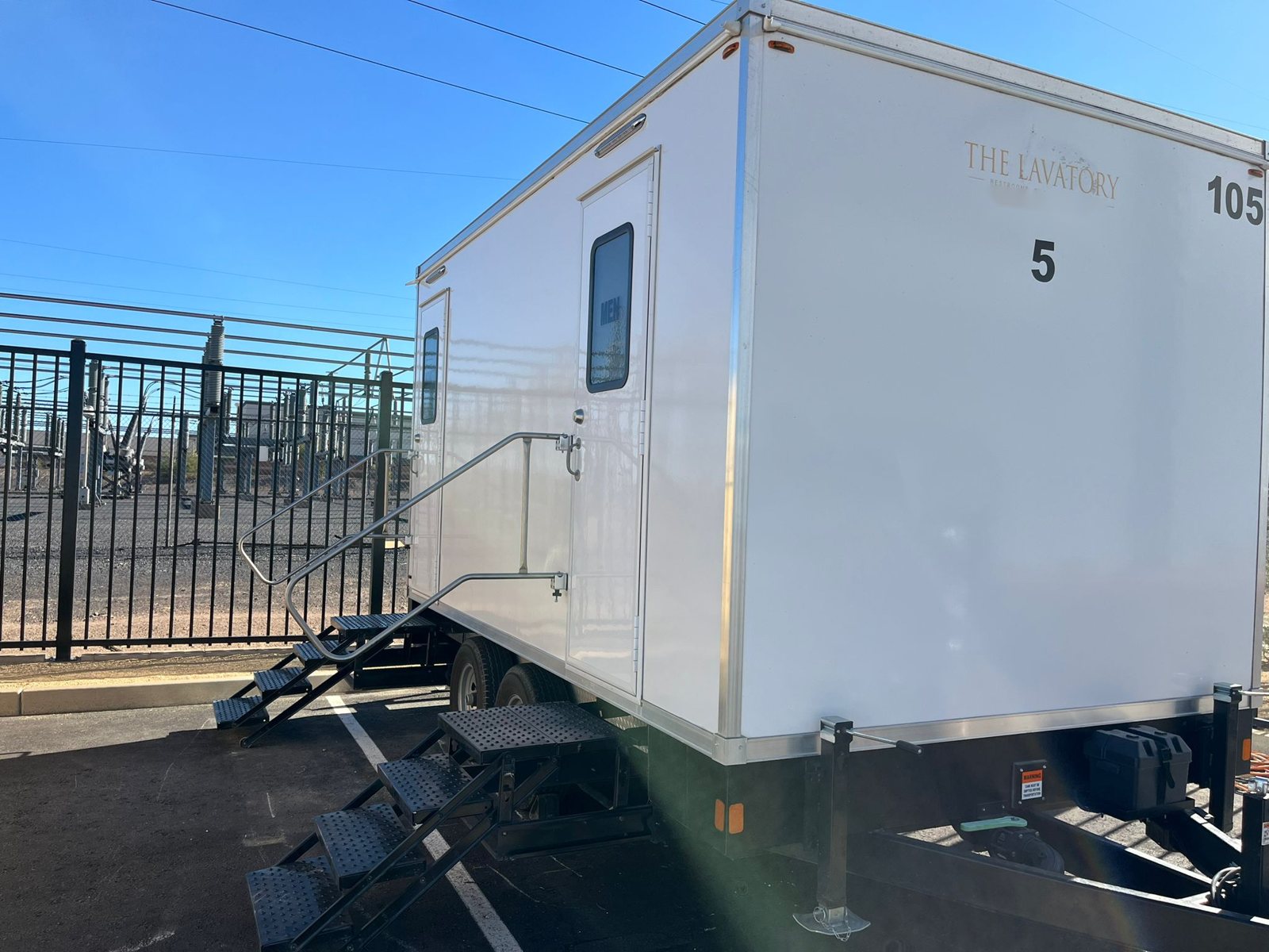A Lavatory 5-Station restroom trailer parked on a paved lot beside a fenced electrical substation on a sunny day