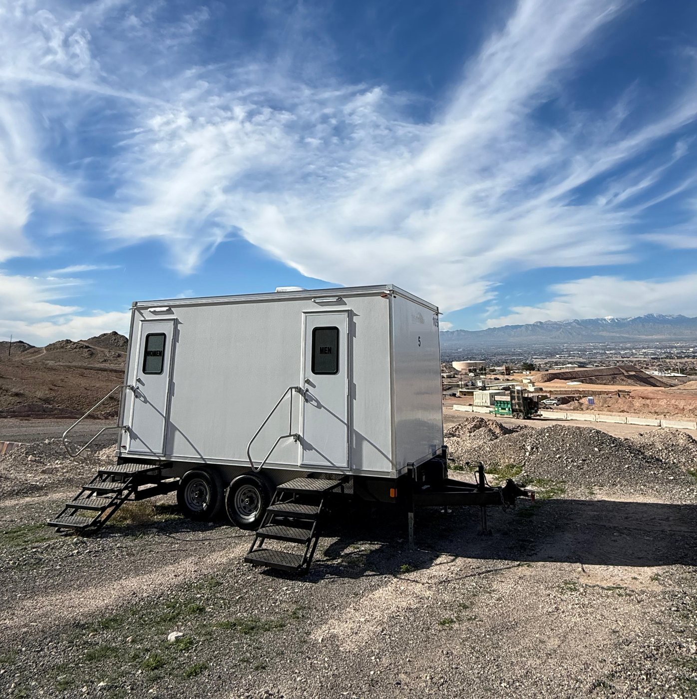 A Lavatory 2-Station restroom trailer set on a gravel desert lot under a wide blue sky with mountains in the distance