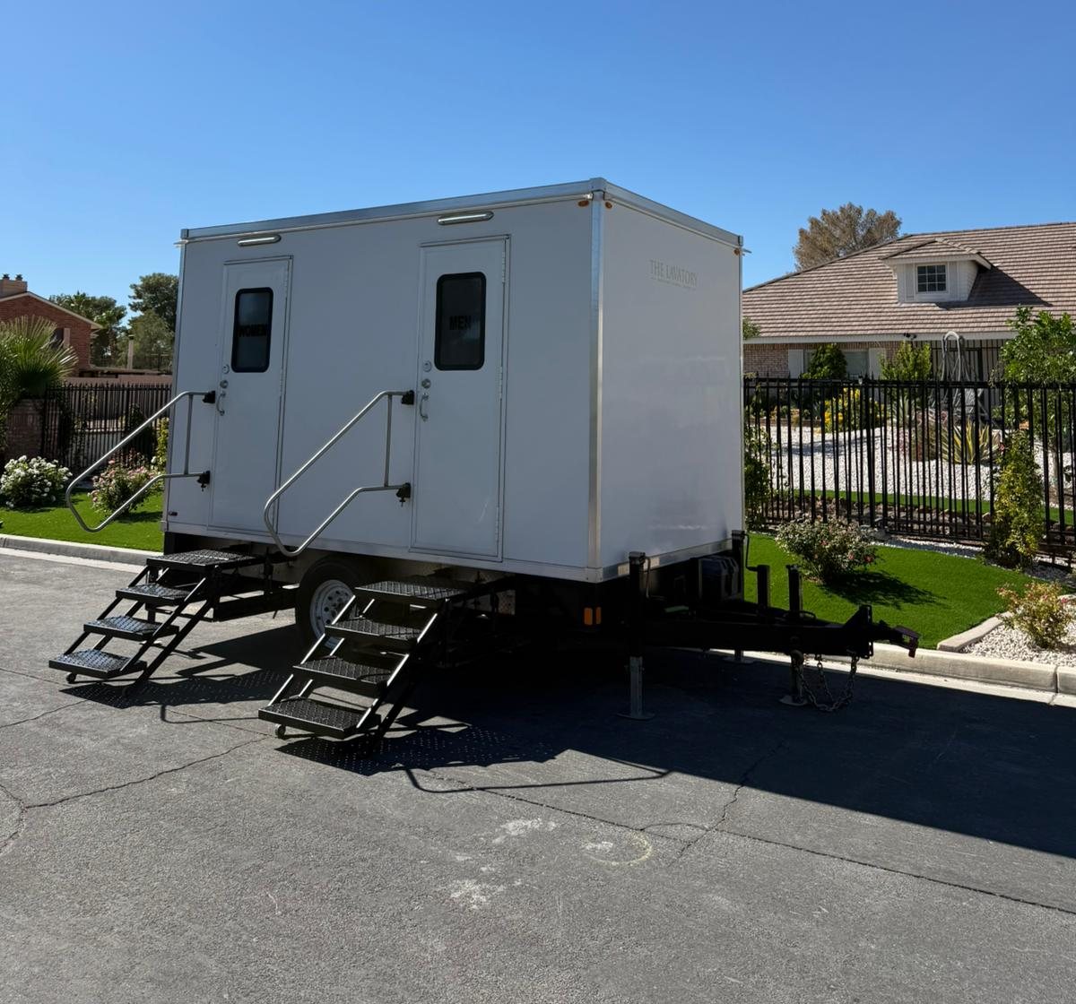 A Lavatory 2-Station restroom trailer parked at the curb beside an iron-fenced home with desert rock landscaping and a green lawn