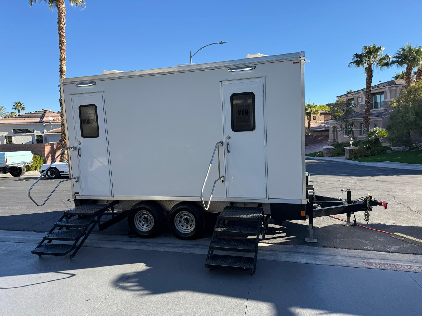 A Lavatory 2-Station restroom trailer with steel stairs parked on a quiet residential street lined with palm trees and stucco homes