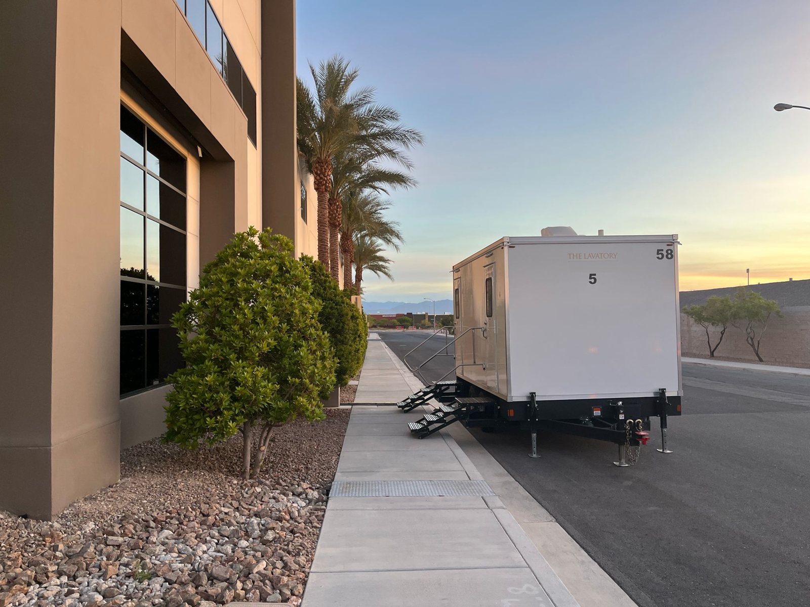 A Lavatory 5-Station restroom trailer parked at the curb beside an office building with palm trees and rock landscaping at sunrise