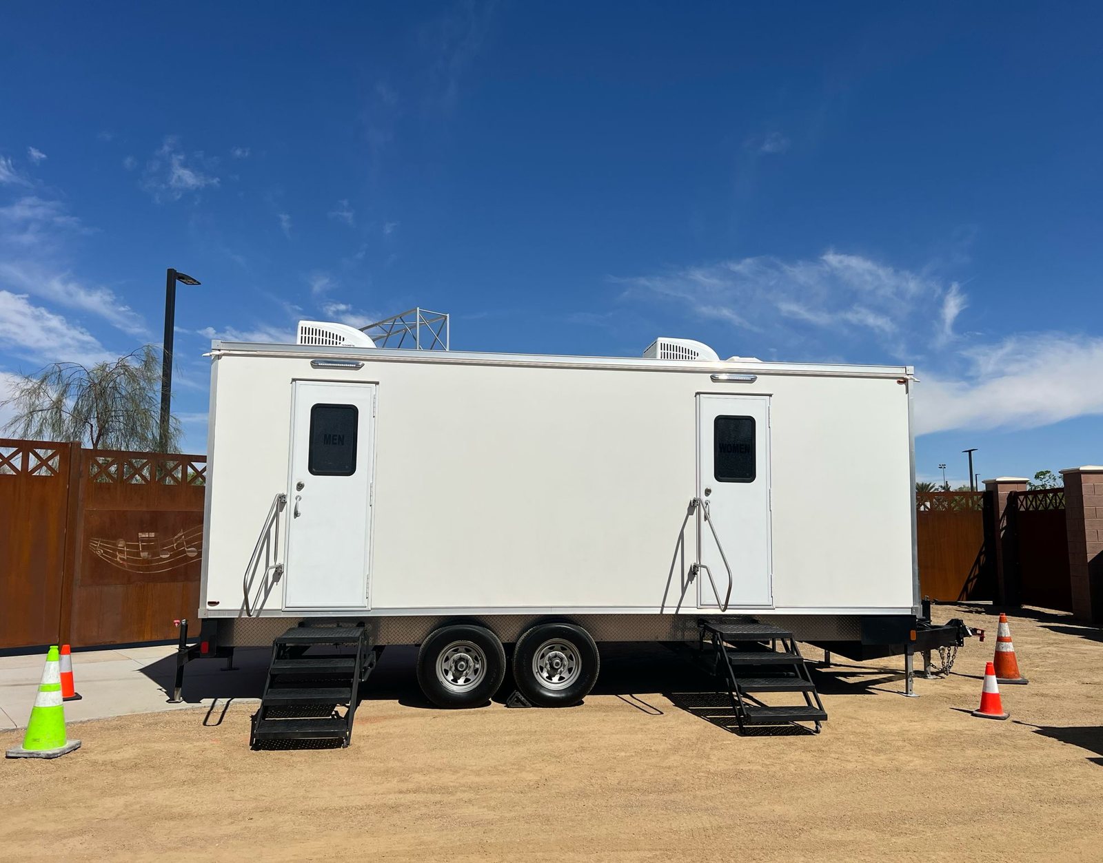 A white Lavatory restroom trailer with men's and women's doors, steel stairs and two rooftop AC units, parked on a desert dirt lot beside a rust-colored metal fence under a deep blue sky