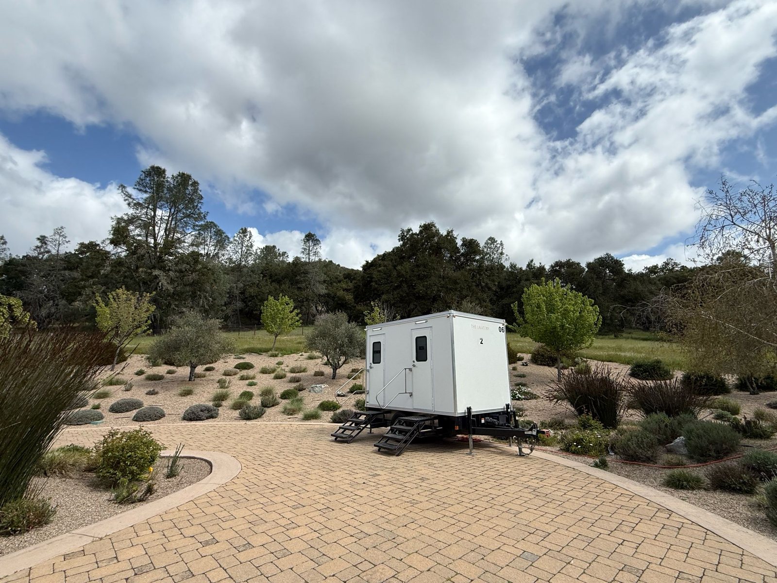 A Lavatory 2-Station restroom trailer parked on a paver courtyard at a landscaped garden venue with drought-tolerant plants and trees