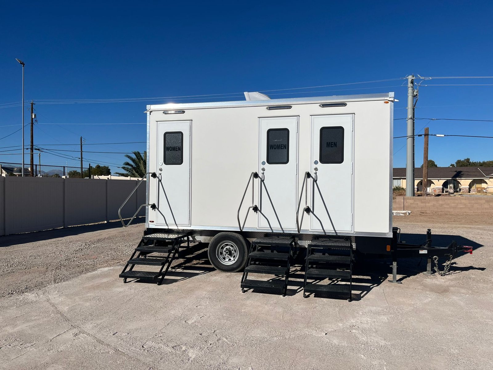 A Lavatory 3-Station restroom trailer with two women's doors and a men's door set up on a gravel lot under a clear blue sky
