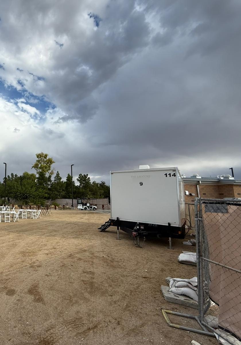 A Lavatory 9-Station restroom trailer set at the edge of a dirt event field with rows of white folding chairs and tables under storm clouds