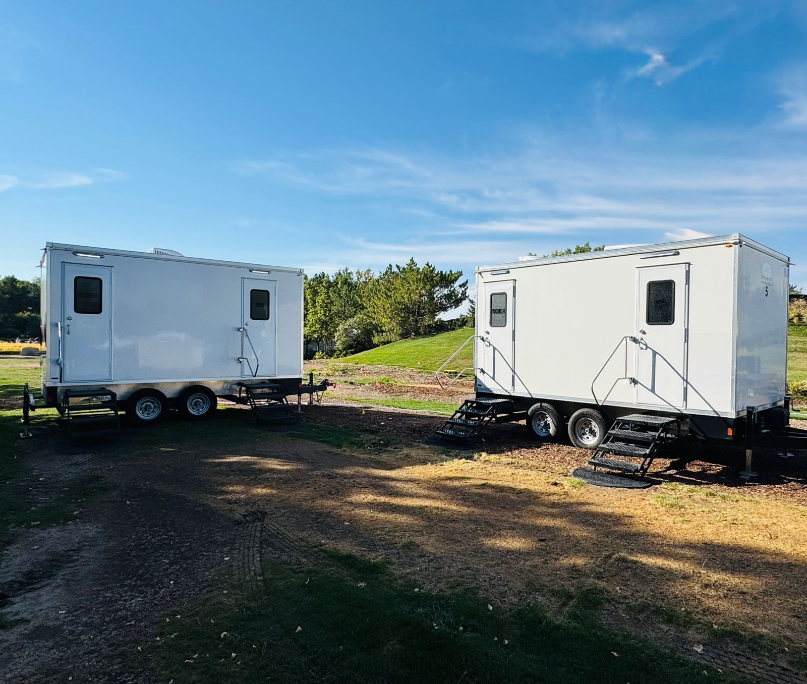 Two Lavatory restroom trailers with steel stairs set side by side on a park lawn for a large outdoor event