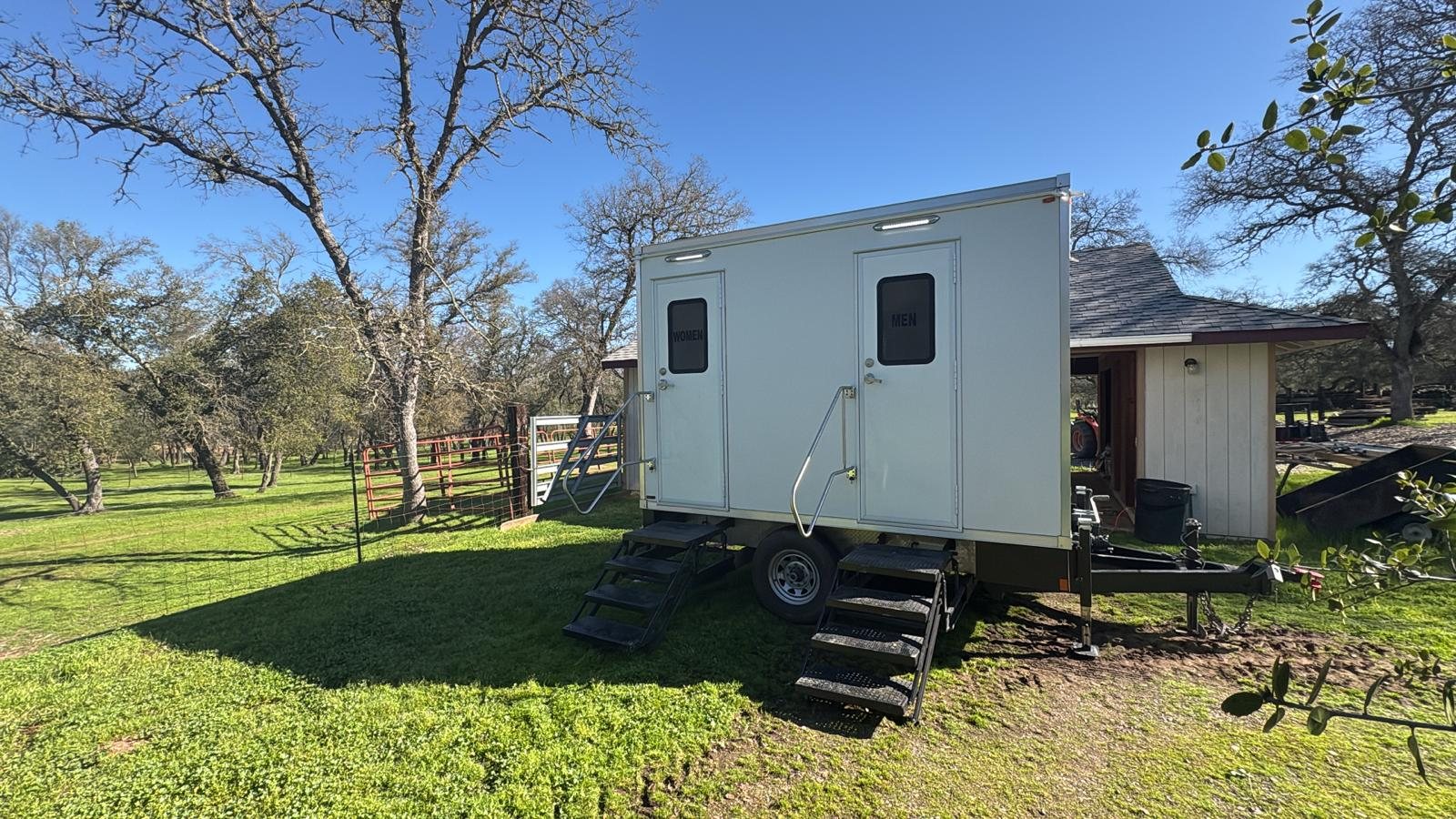 A Lavatory 2-Station restroom trailer on green grass under bare oak trees beside a ranch barn and corral
