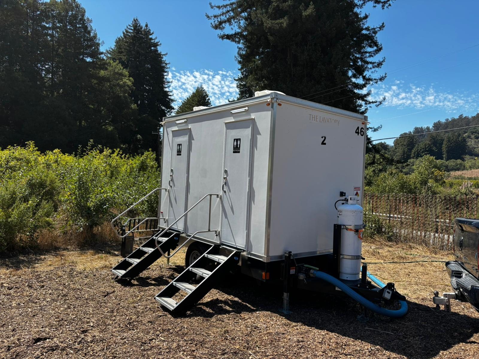 A Lavatory 2-Station restroom trailer on wood chips under tall evergreens with a propane tank at the hitch