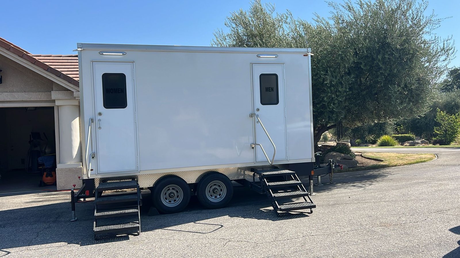 A Lavatory two-door restroom trailer parked in a home driveway beside the garage under an olive tree