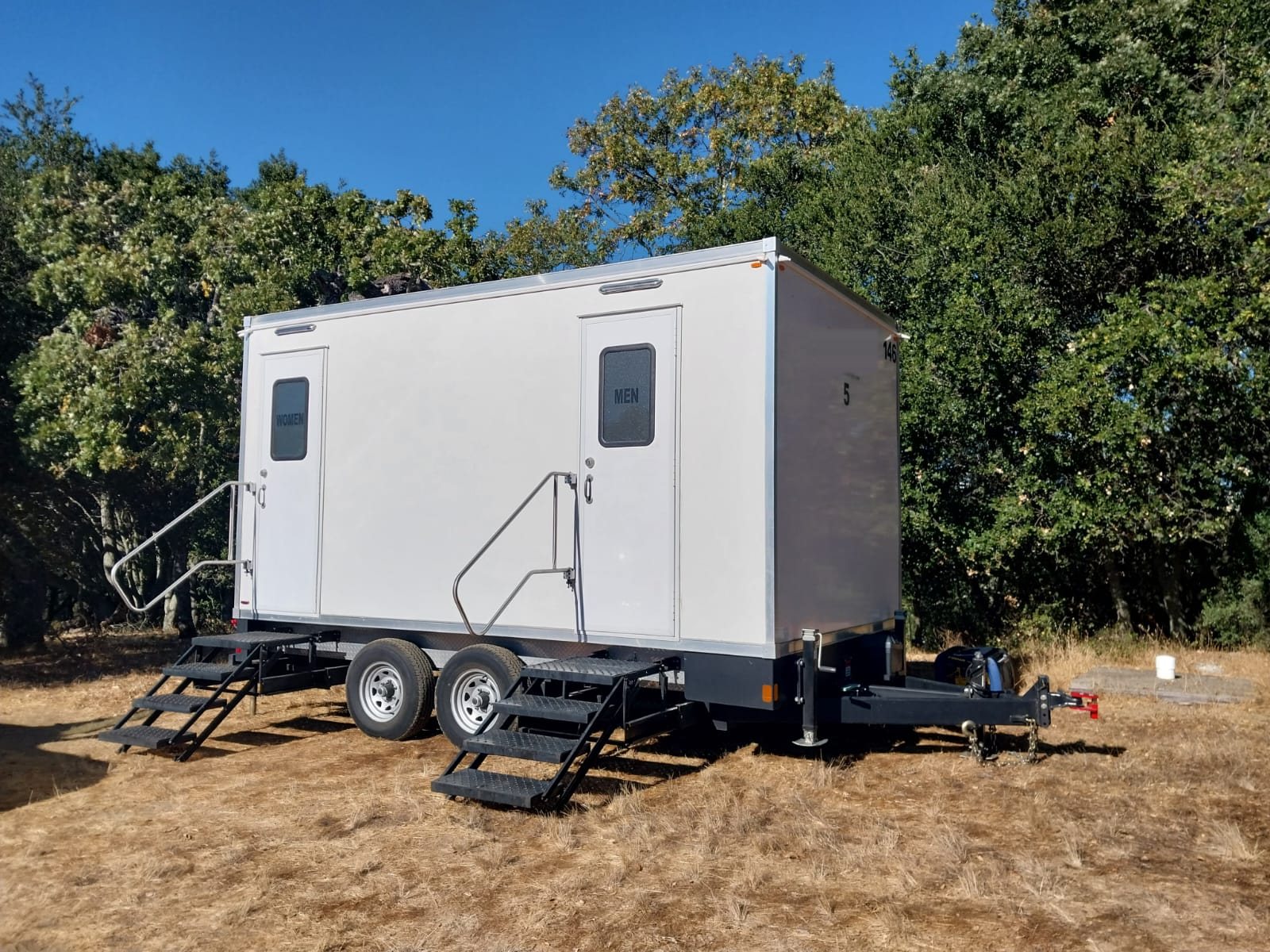 A Lavatory luxury restroom trailer with women's and men's doors set on dry grass in front of tall oak trees