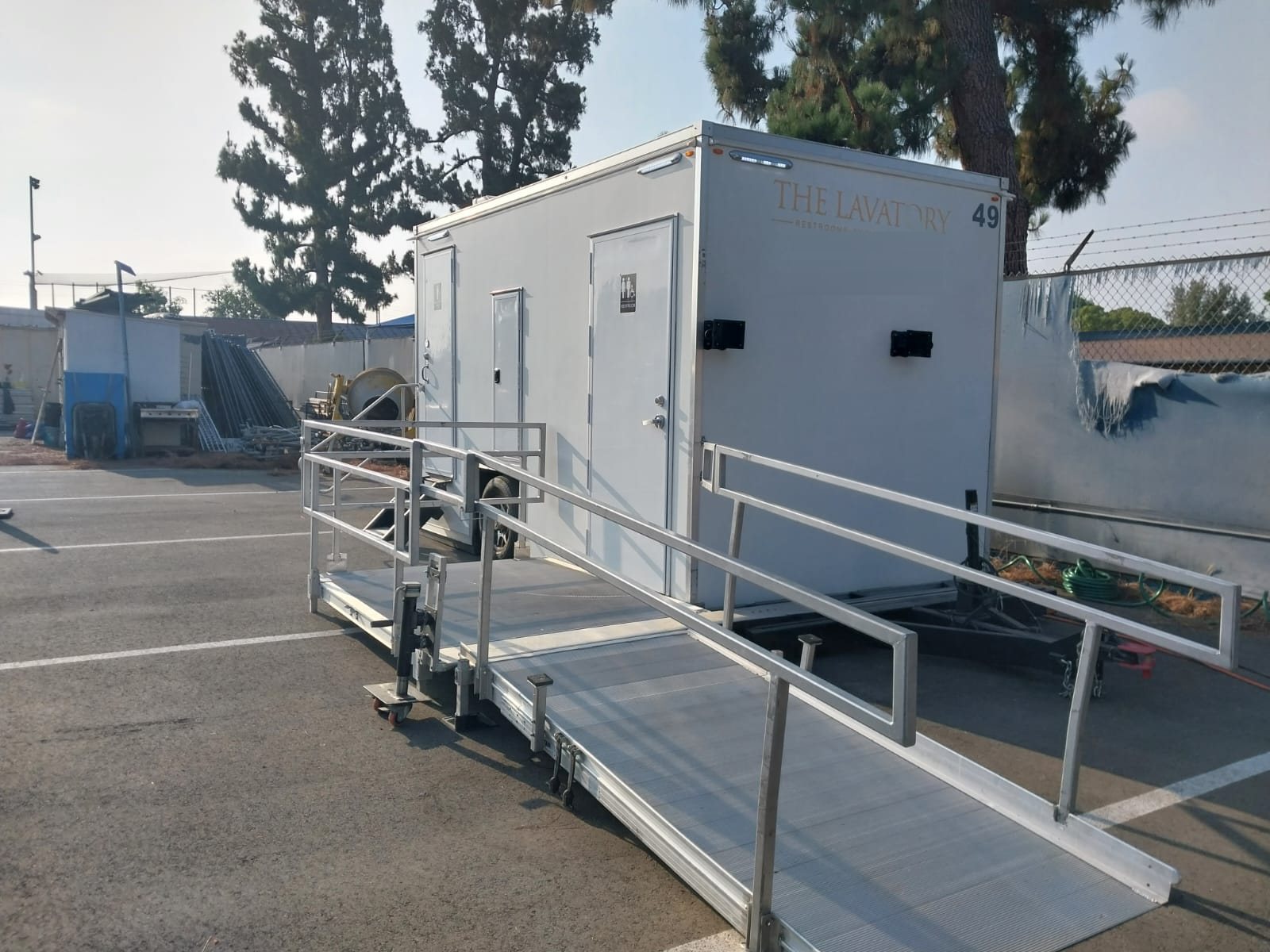 A Lavatory ADA restroom trailer with a long aluminum ramp and handrails in a paved parking lot beside a fenced yard