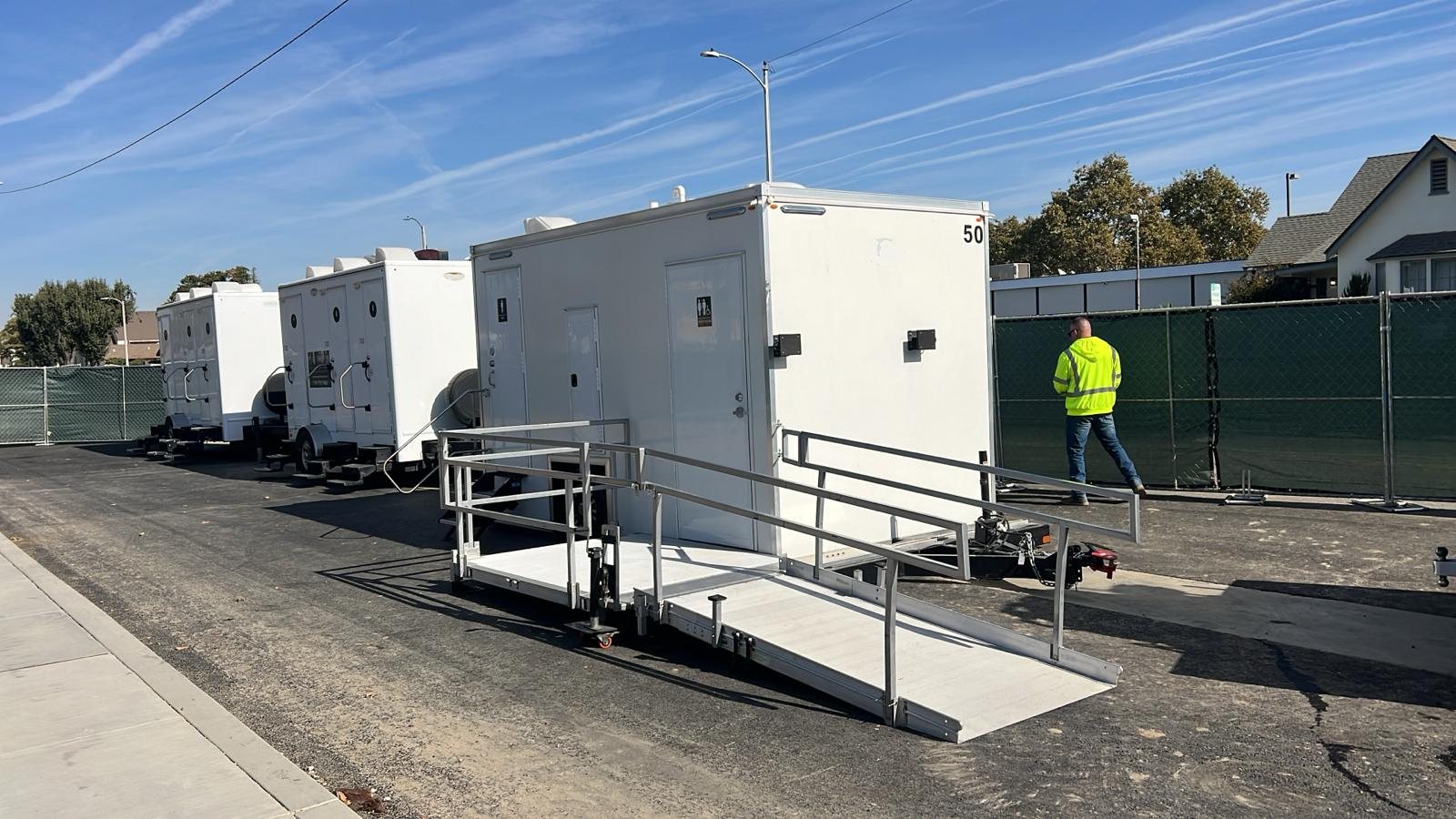A worker in a safety vest walks past a Lavatory ADA restroom trailer with its ramp and handrails set, with more restroom trailers lined up behind it