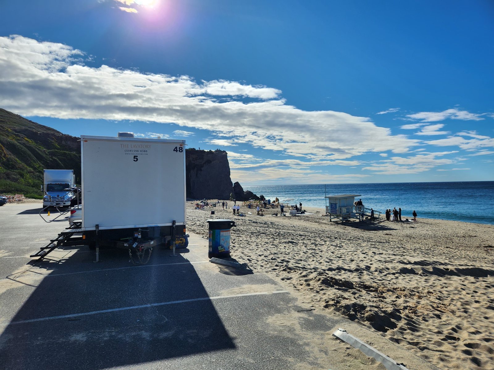 A Lavatory restroom trailer parked at the edge of a beach parking lot, with beachgoers and a lifeguard tower on the sand behind it