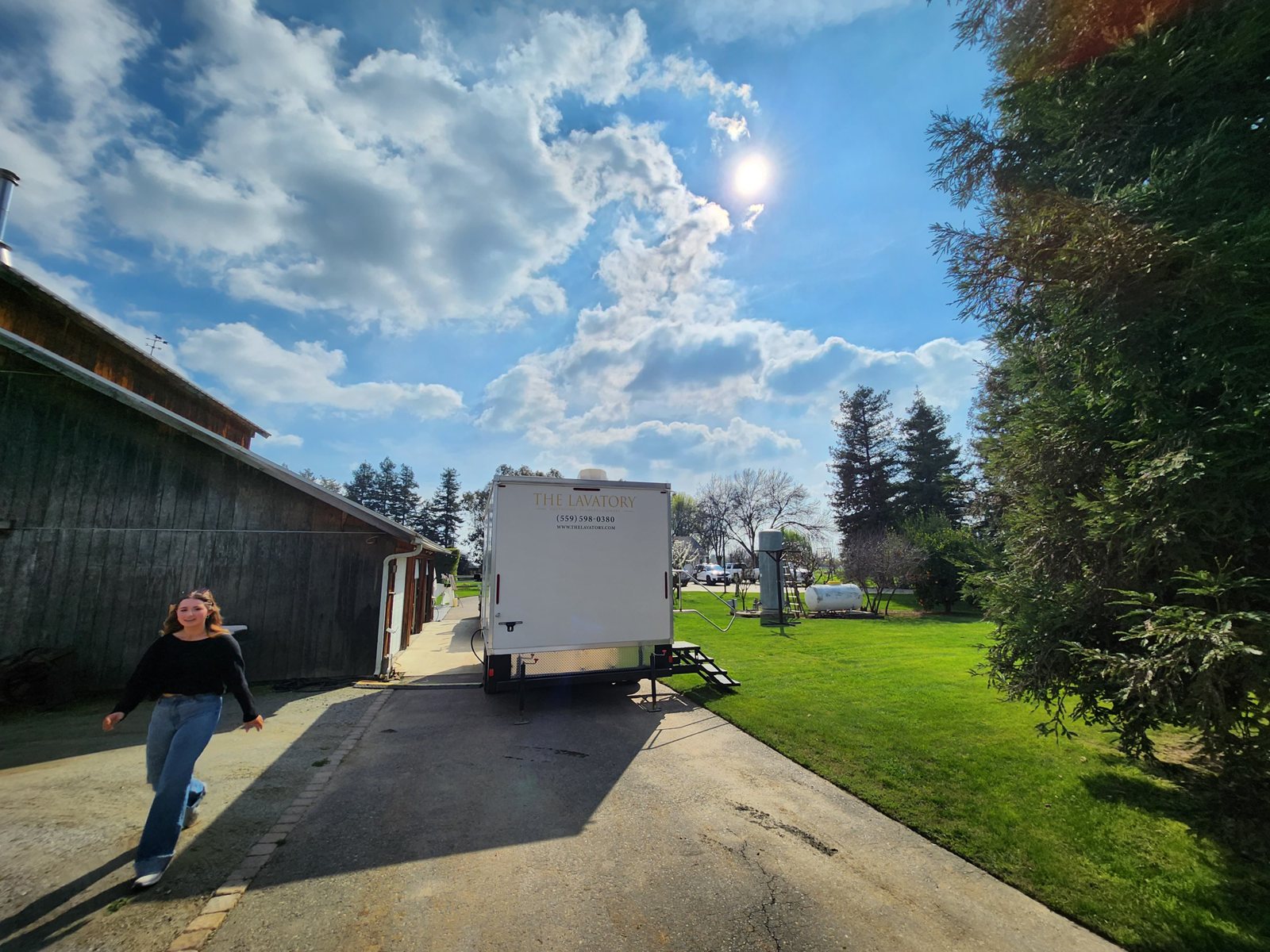 A woman walks up the drive from a Lavatory restroom trailer parked beside a barn at a rural venue
